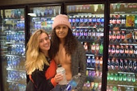 two women standing in front of a refrigerator with soda bottles